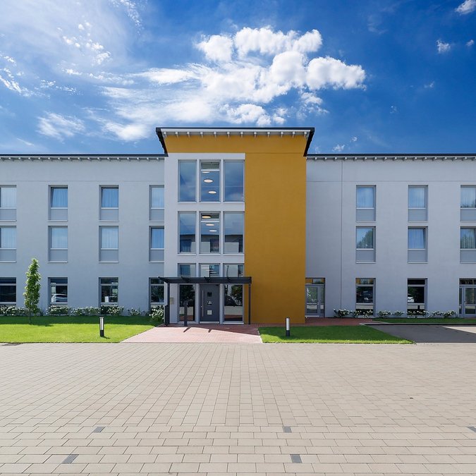 Median Hotel in Lehrte – wir geben Raum Modernes dreistöckiges Bürogebäude mit gelbem Mittelteil und Parkplatz