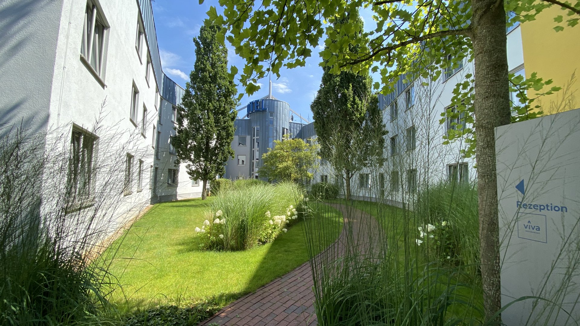 Grüner Innenhof mit Weg zwischen weißen Gebäuden und Bäumen unter blauem Himmel
