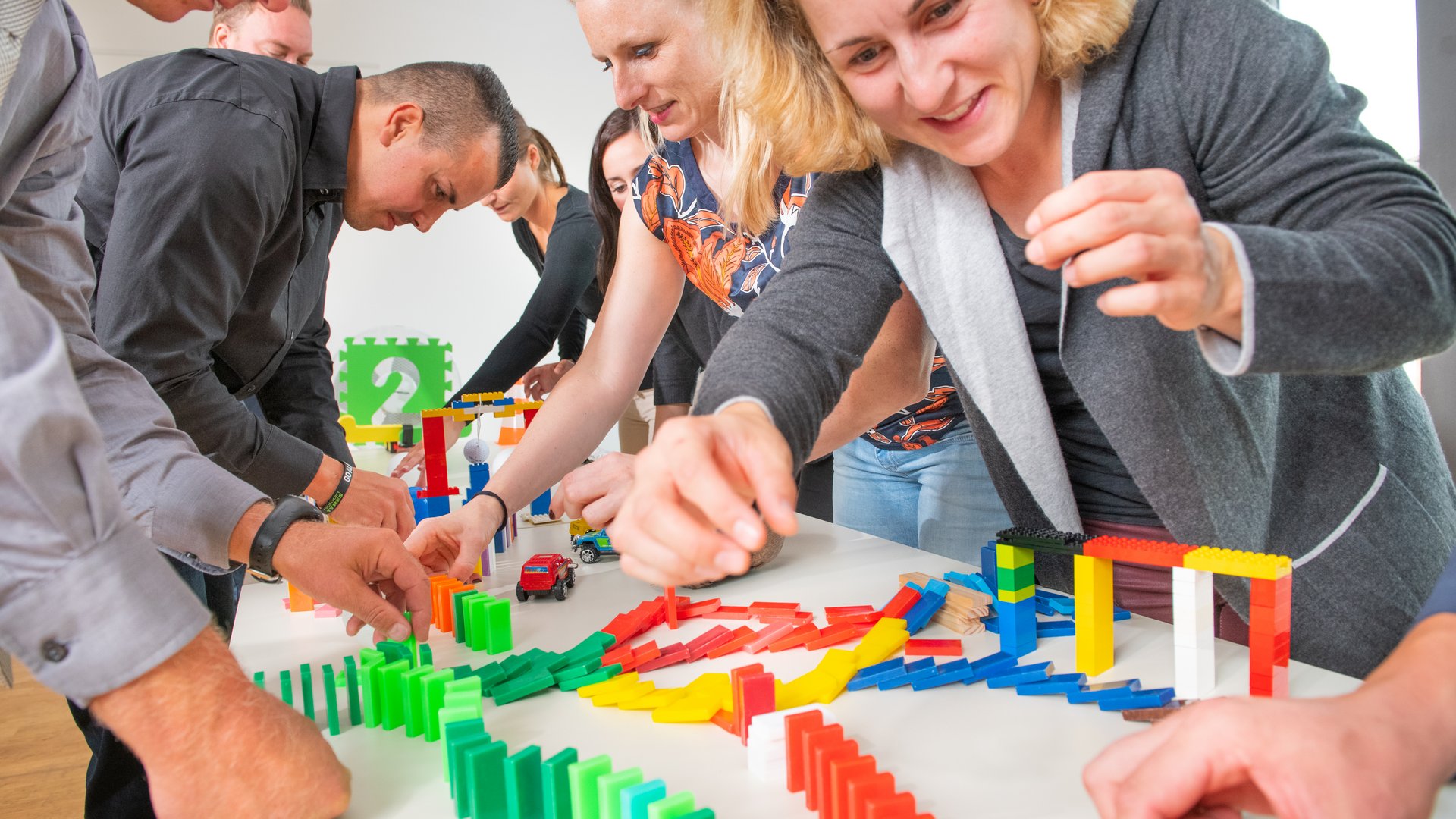 Teambuilding in Hannover und Umgebung Gruppe von Erwachsenen baut bunte Dominosteine und Lego-Struktur auf einem Tisch auf