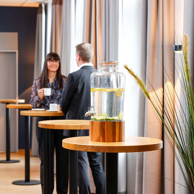 Ihr Tagungshotel in Hannover Glasbehälter mit Zitronenwasser und zwei Menschen im Gespräch im Hintergrund