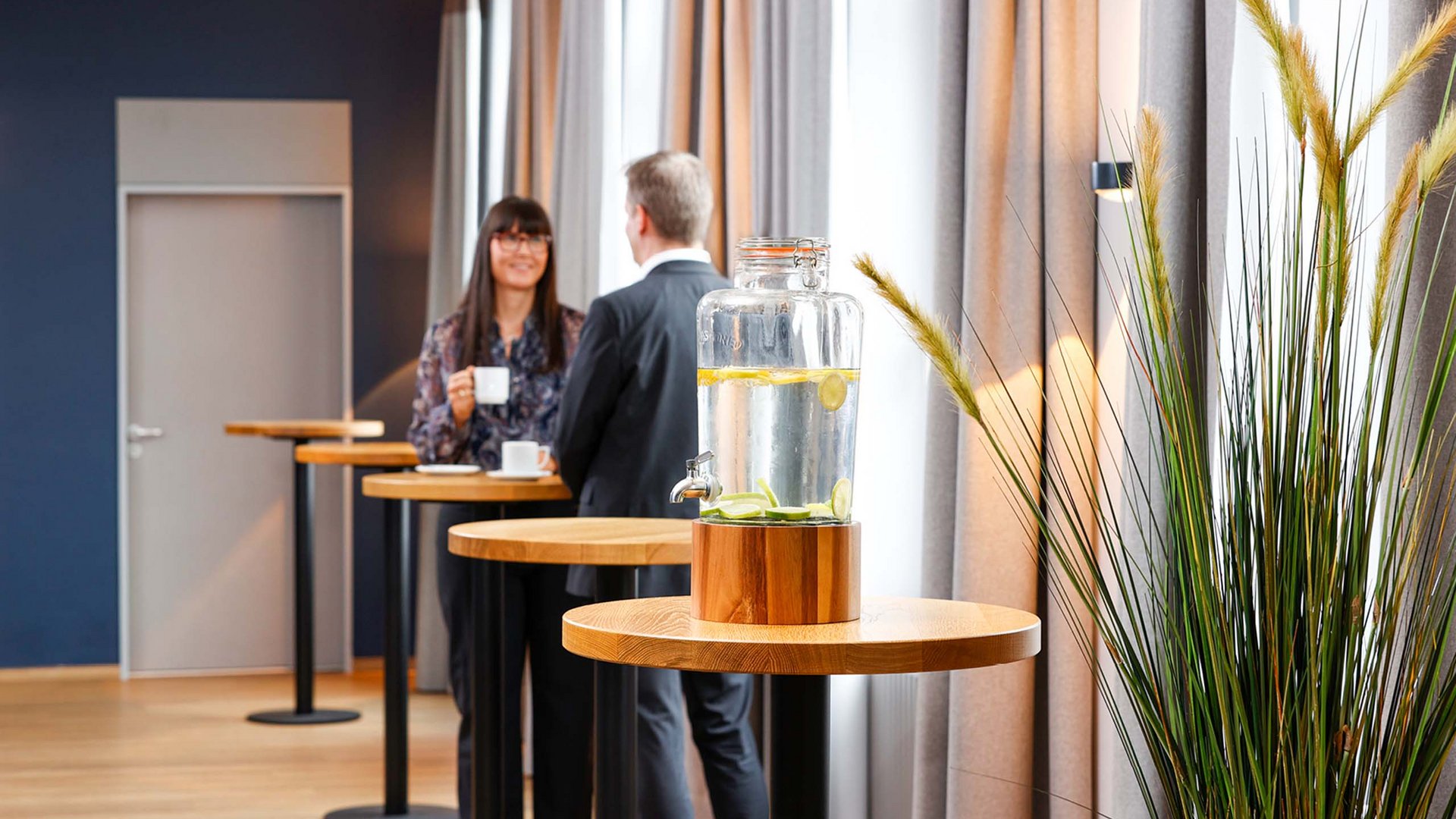 Der perfekte Veranstaltungsort für Tagungen und Konferenzen Glasbehälter mit Zitronenwasser und zwei Menschen im Gespräch im Hintergrund