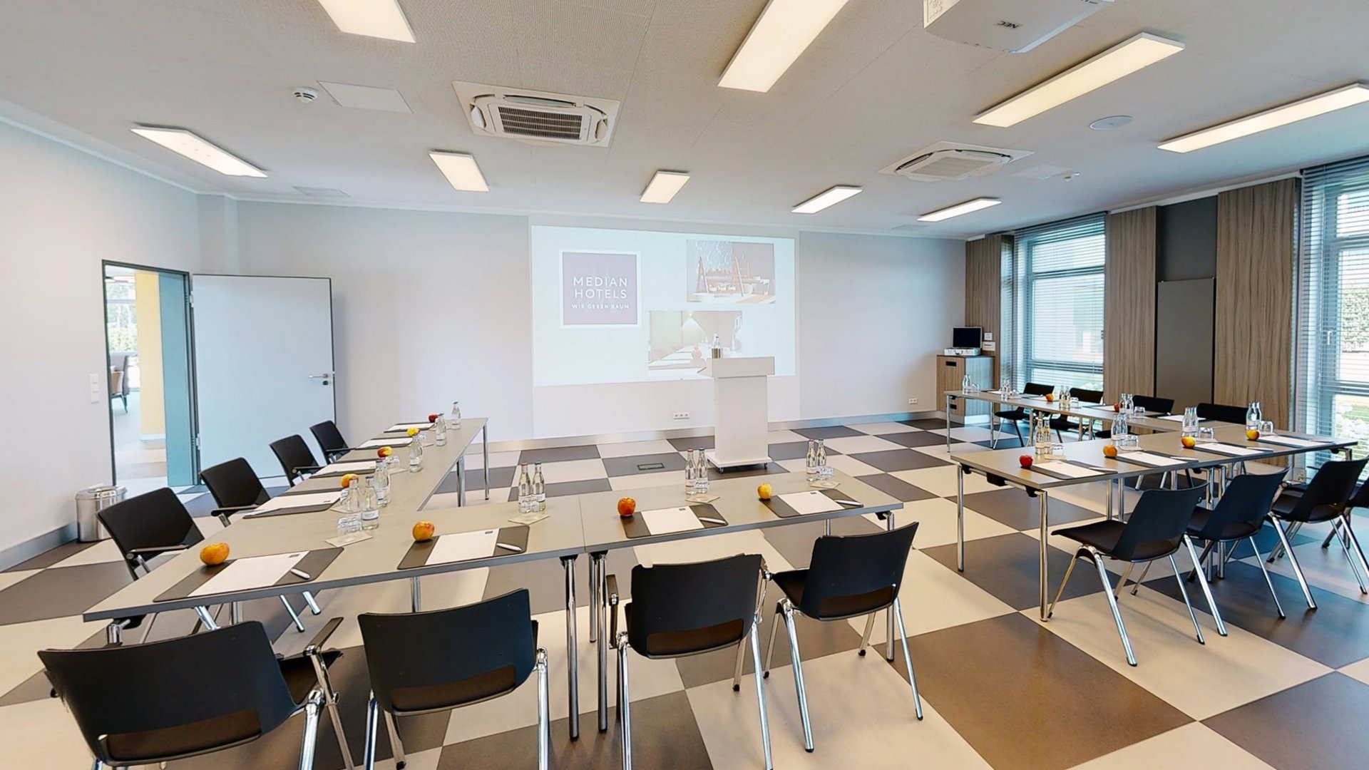 Raum 4 Seminarraum mit Tischen, Stühlen, Wasserflaschen und Äpfeln, Präsentation an der Wand