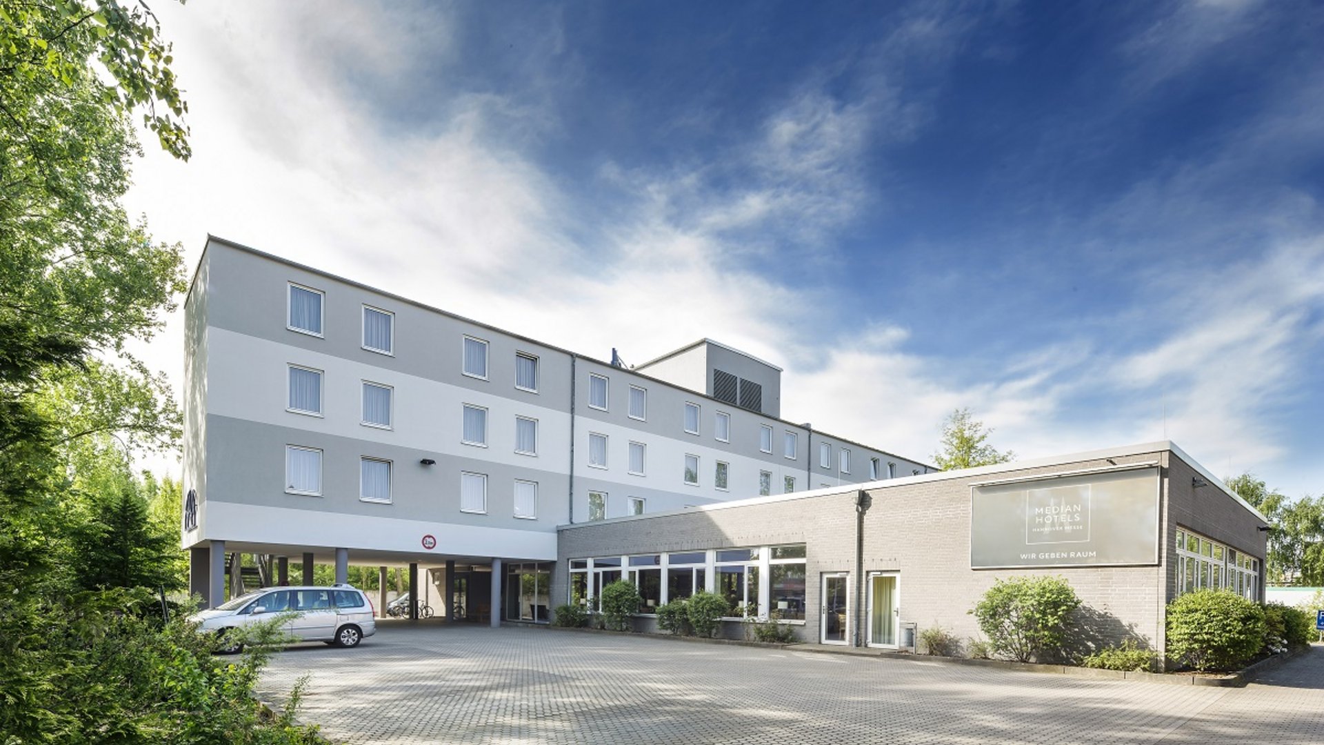 Welcome to the Median Hotel Hannover Messe! Exterior view of a modern hotel with parking lot and surrounding greenery