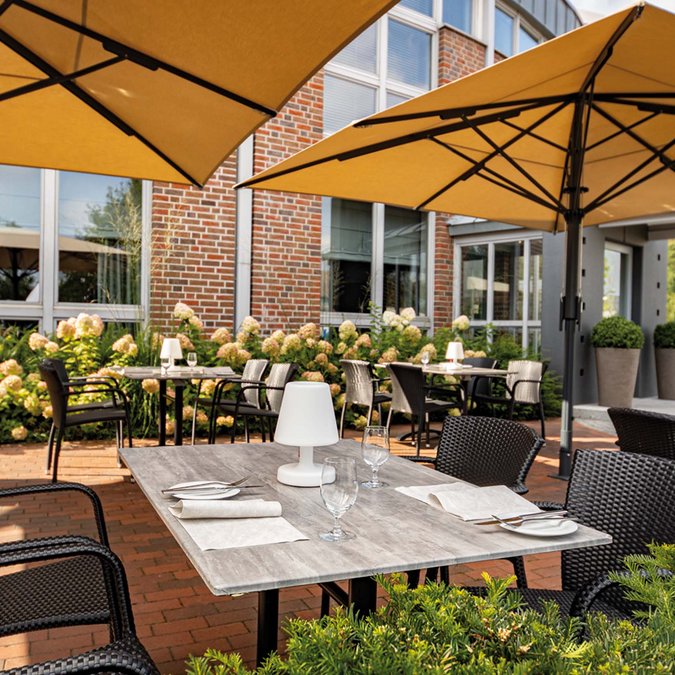 Median Hotel in Lehrte – we provide space Outdoor terrace with tables, chairs, and yellow umbrellas in front of brick building