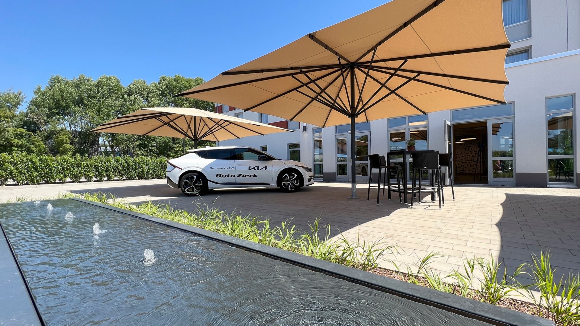 Ihr autarker und exklusiver Tagungsbereich: Ihr Hotel im Hotel Weißer Kia EV6 vor Gebäude mit Sonnenschirmen und Wasserbrunnen