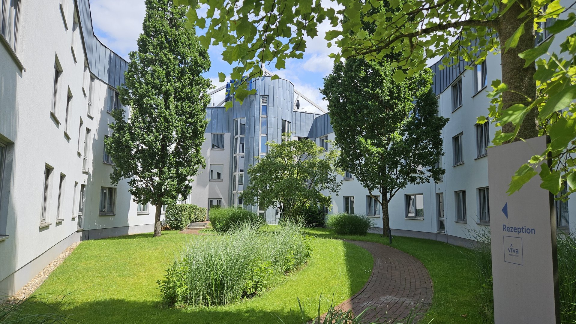 Median Hotel Hannover Lehrte Innenhof mit grünen Bäumen und Gebäuden umgeben bei Sonnenschein