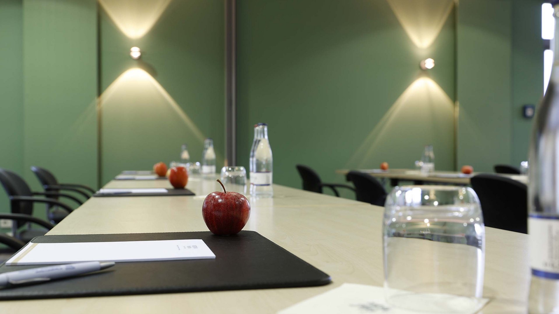 Raum 20+21 Konferenztisch mit Äpfeln, Wasserflaschen und Schreibutensilien in einem Raum mit grünen Wänden